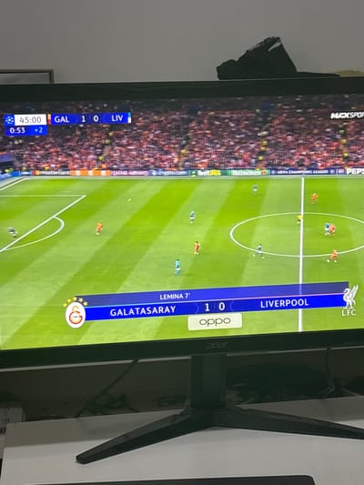 An Acer flat-screen monitor, situated in a room in Krems an der Donau, Austria, displays a live football match. The screen shows a UEFA Champions League game where Galatasaray (GAL) is leading Liverpool (LIV) 1-0. The top-left scoreboard indicates the time as 45:00 with 0:53 seconds into 2 minutes of added time. A bottom graphic highlights "LEMINA 7'" as the scorer, alongside the team names and logos, with an "OPPO" sponsorship visible. The football pitch is green with white markings, and several players in red and light blue/turquoise kits are visible. The stadium stands behind the pitch are densely packed with spectators. Pitchside advertisements include Mastercard, Qatar Airways, crypto.com, bet365, Heineken, UEFA Champions League, and Pepsi. The monitor itself has a black bezel and a black, three-pronged stand, resting on a light-colored surface. A dark, unidentifiable object is positioned on top of the monitor.