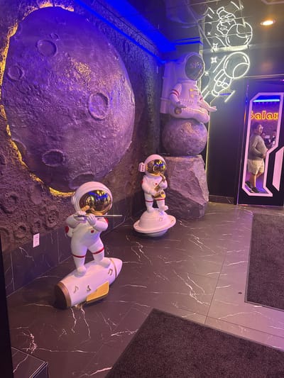 The image shows an indoor scene with a space-themed decor. In the foreground, a statue of an astronaut playing a flute stands on a rocket-shaped base, with musical notes depicted on the rocket. To the right and further back, another astronaut statue is depicted playing a saxophone, standing on a circular platform. Behind these statues, a large, textured sphere resembling a planet dominates the left side of the image. The wall behind the planet is textured and illuminated with purple and blue lights. In the background, a neon sign of an astronaut is visible on the ceiling, reflected in a mirror. A doorway on the right side of the image reveals a person standing in what appears to be a reception area, with a sign that reads "Galaxy" above them. The floor is covered in dark marble tiles with white veining. Two dark gray mats are visible on the floor. The overall impression is of a playful and imaginative entertainment or recreational space. The location is specified as Des Plaines, United States of America.