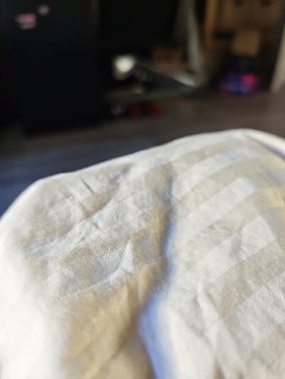 A close-up, slightly blurred photograph shows a beige fabric with a subtle pinstripe pattern. The fabric appears to be part of a comforter or bedding, with gentle folds and creases visible. In the background, out of focus, is a dark, indistinct shape that could be a piece of furniture, and further back, a hint of a room with some colorful objects, possibly toys or decorative items, suggesting an indoor environment, likely a residential setting in Вилла Фортуна Кп, Russia.