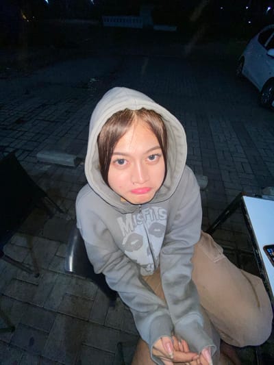 A young woman with long brown hair and prominent eyelashes is seated outdoors at night in what appears to be a paved area with a brick-like pattern. She is wearing a light gray hooded sweatshirt with a graphic design and brown pants. Her facial expression is one of mild displeasure or poutiness. In the background, a white car is visible, and the lighting suggests it is evening or nighttime. The scene is located in Mekar Bakti, Indonesia.