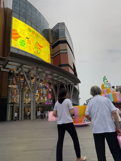 In the Haidian District of China, a wide shot captures the exterior of a modern shopping mall under an overcast sky. The mall features a curved facade with large glass windows and distinct brown and beige paneling. Dominating the left side of the image is a massive, bright yellow LED screen displaying colorful graphics and Chinese text, promoting an event. The screen's content includes text like "DREAM SEEK" and references to anniversaries and discounts.
In front of the mall's entrance, a series of large, metal support beams create an architectural canopy over the glass doors. Decorative lights, in various colors and patterns, are visible through the glass entrance, forming intricate shapes and outlines.
Several people are visible walking across a paved plaza in front of the mall. Two women are in the foreground, with their backs to the camera. The woman on the left wears a striped shirt and black pants, while the woman on the right has on a white shirt with a dark floral pattern and black pants. Further back, other individuals can be seen entering and exiting the mall, as well as strolling through the plaza.
To the right of the mall's entrance, a large, colorful inflatable sculpture resembling a stack of oversized cups or tubes is prominently displayed. The sculpture is predominantly pink, yellow, and green, and the words "DREAM SEEK" are visible on some of its sections. A pink object, possibly part of the sculpture or a separate display, is also visible on the ground. The ground itself is paved with large, rectangular tiles.