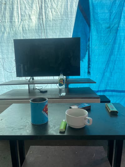 A television is mounted on a stand, behind which are two shelves holding a bottle of Lipton iced tea. In front of the television, on a dark table, are a blue can with a Superman logo, a pack of cigarettes, a white mug filled with a dark liquid, a green lighter, and a gold-colored lighter. The scene is set against a backdrop of a light-colored tarp on the left and a blue tarp on the right. The overall impression is of an outdoor or makeshift living space, possibly in a rural or temporary setting. The image does not provide specific geographic markers to definitively place it in Tsalyk, Russia, but the presence of these everyday items suggests a domestic or personal space.