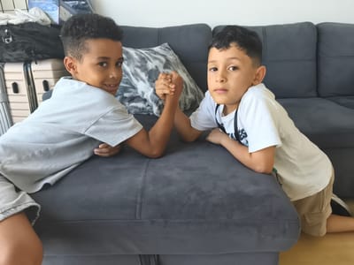 Two young boys are arm wrestling on a dark grey couch. The boy on the left has curly dark hair and is wearing a light grey t-shirt and shorts. He is looking towards the camera with a slight smile. The boy on the right has dark, straight hair and is wearing a white t-shirt with a black graphic and tan shorts. He is looking directly at the camera with a neutral expression. Their hands are clasped in a strong grip as they compete. Behind them, a textured grey pillow is visible on the couch, and further back, suitcases and other items are stacked, suggesting a travel or moving context. The setting appears to be indoors, likely a living room, with a wooden floor visible in the foreground. The lighting suggests daytime.