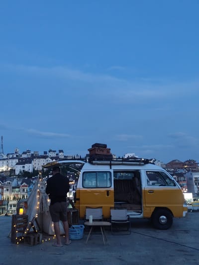 A man stands near a yellow and white van parked on a flat, possibly rooftop, surface in what appears to be the city of Đà Lạt. The van's side door is open, revealing a cluttered interior. A makeshift tent-like structure, adorned with fairy lights, stands adjacent to the van. The man, facing the tent, wears a dark jacket and shorts. Two folding chairs and a small table are set up near the van. In the background, there is a view of the city's architecture with closely built, multi-story buildings and a tower. The sky is dark, indicating twilight or evening. A blue plastic laundry basket is near the tent. Suitcases are strapped to the roof of the van.