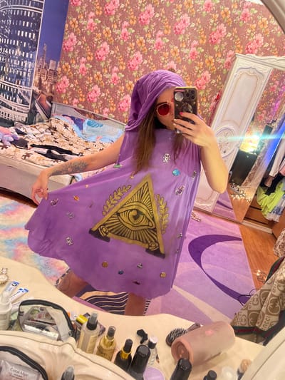 A person is standing in front of a mirror, taking a selfie. They are wearing a purple hooded dress with an all-seeing eye and celestial motifs. The background appears to be a bedroom with floral wallpaper, a cluttered bed, and a large wardrobe. There are various items on a dresser in the foreground, including makeup, a brush, and a speaker. The image does not provide any information about the city of Krasnoe-na-Volge, Russia.