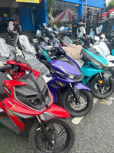 The image depicts a street-level view of a motorcycle dealership in Alor Setar, Malaysia. The dealership's exterior is visible, featuring blue walls and a prominent yellow awning. A row of new motorcycles, some covered in protective plastic, are parked on the asphalt. The motorcycles are of various colors, including red, purple, and turquoise.

In the foreground, a red motorcycle with "MODA AERO E" branding is prominently displayed. Behind it, a purple motorcycle and a turquoise motorcycle with "MORBID\" branding are visible. In the background, several individuals are present. Two men are seen near a truck, possibly unloading or preparing motorcycles. Another person, wearing a hijab and a black jacket, is walking with a man in a black t-shirt near the dealership entrance. The environment suggests a commercial area, with a focus on motorcycle sales and services. The asphalt surface appears wet, indicating recent rain.