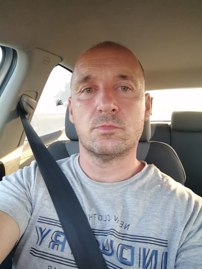 A man in a grey t-shirt with a dark seatbelt across his chest is seated in the driver's seat of a car. He has short brown hair and stubble. The interior of the car is visible, including the dashboard and the headrest of the passenger seat. The image appears to have been taken in natural daylight.