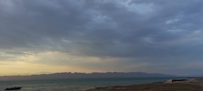 The image shows a body of water with a distant mountainous shoreline under a cloudy sky. The water is a turquoise green, and the shore is rocky and appears to be composed of sand and pebbles. Several thin, vertical poles, likely fishing rods, are scattered along the water's edge. In the foreground, a small, dark boat is partially submerged near the left edge of the frame. The sky is filled with heavy, dark gray clouds, with patches of lighter, yellowish light breaking through near the horizon. The mountains in the distance are silhouetted against the sky, with layered peaks stretching across the frame. The overall scene suggests a natural landscape, possibly at dusk or during overcast weather. No people are clearly visible in the image.