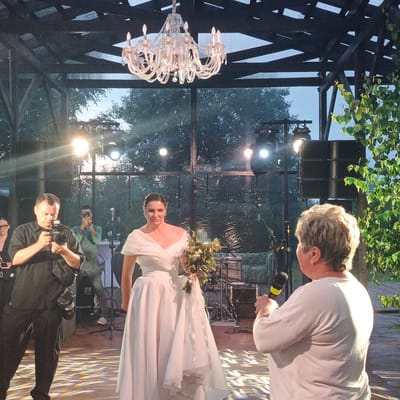 The image captures an outdoor event, possibly a wedding reception, at the "Svetloye" restaurant complex in Svetloye, Russia. In the foreground, a woman in a white wedding dress stands holding a bouquet of greenery. To her left, a man in a black shirt and pants is photographing her with a professional camera. Behind the bride, another man is seen taking a photo with his phone. In the mid-ground, a speaker system and stage lighting are visible. The background features trees and the sky, indicating an evening or late afternoon setting. The overall atmosphere appears festive, with a large crystal chandelier hanging from the structure above.