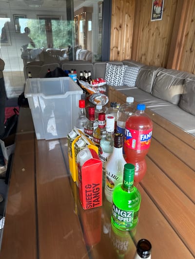 The scene depicts a semi-enclosed outdoor or conservatory space, likely a patio or decking area, in Bubwith, United Kingdom. A large wooden table with a reflective glass surface occupies the foreground and mid-ground, laden with beverages and snacks.
On the table, a prominent array of alcoholic spirits includes multiple bottles of Smirnoff vodka, Bacardi rum, Malibu, and a green bottle of Tang Sour Apple liqueur. One Smirnoff bottle has a red price tag of "£16.99". Several cartons of juice or mixers, including one labeled "SWEET & TANGY," are positioned alongside. A large two-liter plastic bottle of Fanta with red-orange liquid is also present. A clear, rectangular plastic storage bin sits on the left side of the table. Further back, several dark and amber glass beer bottles are visible, intermingled with opened bags of crisps or snacks.
The background features an L-shaped outdoor sofa furnished with light grey cushions and multiple black-and-white patterned throw pillows. The wall behind the sofa is composed of vertical wooden slats, with a framed poster (partially visible, possibly depicting a cocktail) hanging on it. Large glass panels or windows reflect the interior space, showing the silhouettes of at least two individuals, one standing and one partially visible, suggesting movement within the adjacent area. The setting indicates a prepared environment for a social gathering or casual event.