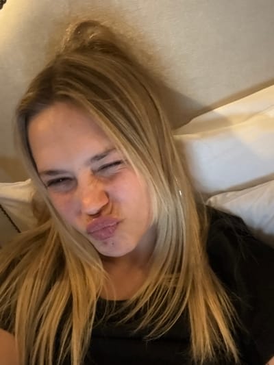 A woman with blonde hair lies on a pillow, posing for a selfie. She has one eye closed and a pouty expression. Her hair is long and slightly tousled, with some strands falling across her face. She is wearing a dark-colored top. The pillow is white and plush, contrasting with the darker tones of her clothing.