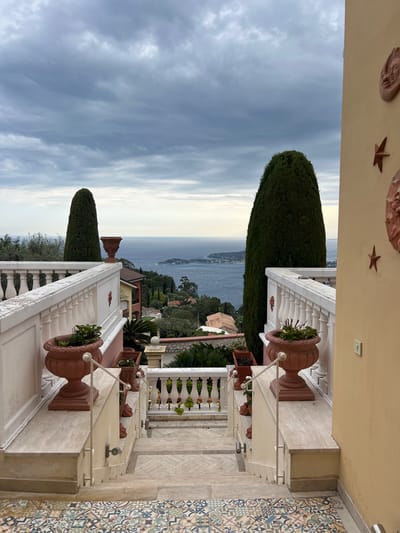 An elevated view captures a serene Mediterranean landscape under an overcast sky. The image looks out from a multi-tiered balcony or terrace towards a vast expanse of deep blue sea and a distant, undulating coastline dotted with buildings and lush green vegetation.

In the immediate foreground, architectural details frame the scene. On the right, a section of the floor features vibrant, geometrically patterned tiles in hues of blue, yellow, red, and white. Adjacent to this, a white balustrade with classical detailing extends across the lower portion of the frame, adorned with ornate, reddish-brown urn-shaped planters holding green foliage. Some planters have decorative white ropes. Above the balustrade, concrete ledges support small spotlights. To the upper right, a light yellow wall or ceiling displays decorative elements, including a terracotta mask-like face and a star-shaped ornament.

Two prominent, tall, conical dark green cypress trees stand vertically, breaking the horizon line and adding a striking natural element to the view. Beyond these, the sea stretches to a faint, brighter horizon line beneath a soft, cloudy sky. Scattered houses and a winding road are visible along the distant shore.

The weather appears cloudy, lending a soft, diffuse light to the daytime scene. No people or activities are visible, emphasizing the tranquility and picturesque quality of the coastal vista. The setting exudes a quiet elegance, typical of the French Riviera, such as Èze. There is no visible text in the image.
