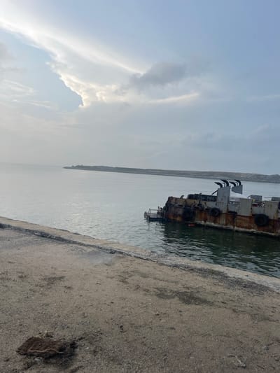 A rusted ship hull sits against a concrete pier in a body of water. The sky is cloudy with patches of blue. The water's surface reflects the sky and the ship. The pier is textured and worn with a section of debris near the lower right corner. The photograph is rotated, presenting a tilted perspective of the scene.