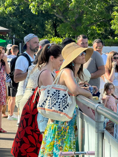 A daytime outdoor scene shows a group of people standing close together in Fentange, Luxembourg. Many of them are looking up and to the left. A woman in the front of the group, facing left, wears a white top and carries a white bag with blue designs. Next to her is a woman in a straw hat, with sunglasses. Above them, others are looking in the same direction, including a man wearing sunglasses. Above that, a child is visible. A metal structure is above the group of people, with metal poles and a white surface. Vegetation, possibly trees, is visible in the background. The people appear to be onlookers at an event.