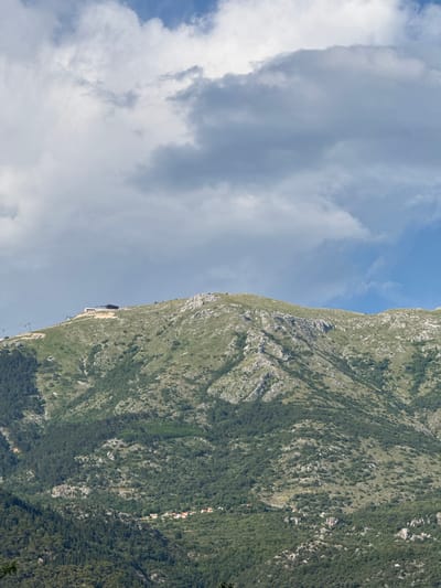 The image shows a view of a steep, green mountainside with a blue sky and clouds above. The mountain is covered in vegetation, with patches of lighter rock visible. There are some small buildings nestled into the hillside. In the distance, a cable car is visible, ascending the mountain. The scene suggests a sunny day with some cloud cover. The overall impression is one of natural beauty and a recreational activity.
