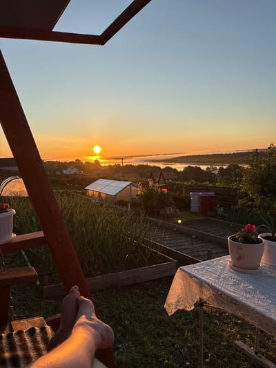 The image depicts a rural landscape in Зеленец, Russia, illuminated by the low, warm light of either sunrise or sunset. In the immediate foreground on the left, the bare feet of an individual are visible, resting on a textured, possibly woven mat, positioned on a dark wooden structure, likely a swing chair or bench. Sunlight directly illuminates the feet. A substantial wooden beam from the same structure frames the upper left and left side of the composition.
To the right in the immediate foreground, a small, collapsible table covered with a white, intricate lace tablecloth is present. Two white potted plants sit on the table; one on the left contains a vibrant red flowering plant, and the pot features a simple drawn figure.
The midground reveals a cultivated garden featuring multiple raised garden beds with tilled soil and young green crops, likely garlic. Two greenhouses are visible: an arched, translucent tunnel greenhouse on the left and a gabled, white-roofed greenhouse on the right, both reflecting significant sunlight. Further back in the garden, two large barrels, one red and one white/grey, are positioned near a wooden fence.
The background presents a village or settlement with scattered houses. Beyond the village, a body of water, possibly a river or lake, is partially obscured by a layer of low-lying mist. The sun, a prominent, bright orange orb, is positioned low on the horizon, directly above the misty water and a distant treeline, casting a strong, golden hue across the entire scene. A utility pole with wires is visible on the right side of the background, extending towards the distant foliage. The sky transitions from a rich orange-yellow near the horizon to a clear, pale blue overhead.