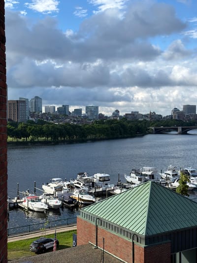 The image shows a waterfront view in Cambridge, Massachusetts, USA. In the foreground, a brick building with a green, sloped roof is visible. Beyond the building, a body of water is present, with several boats docked. Further in the distance, a tree-lined park separates the water from a city skyline featuring various buildings of different heights and architectural styles. A bridge is also visible spanning the water in the mid-ground. The sky is cloudy with patches of blue sky showing through. A police car is parked on a grassy area near the waterfront.