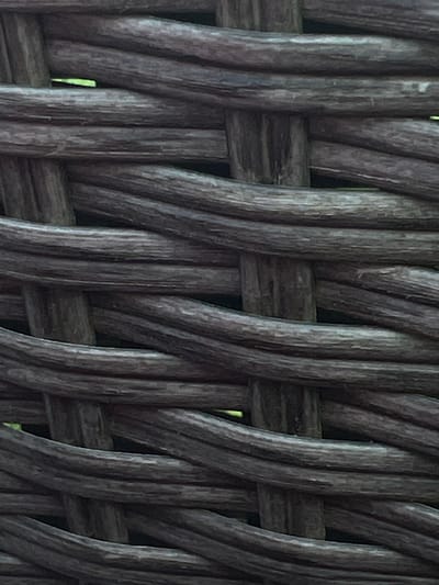 The image displays a close-up view of woven, dark brown material, likely rattan or wicker. The texture is coarse and the individual strands are thick and rounded. The weaving pattern is a simple over-under interlacing. In some of the gaps between the woven strands, a small sliver of green is visible, suggesting an outdoor environment or a backdrop of foliage. The lighting is subdued, creating shadows and highlighting the texture of the material. There are no people or any discernible objects beyond the woven material itself. The image does not contain any specific landmarks or architectural features that would allow for the identification of a precise location within Genemuiden, Netherlands.