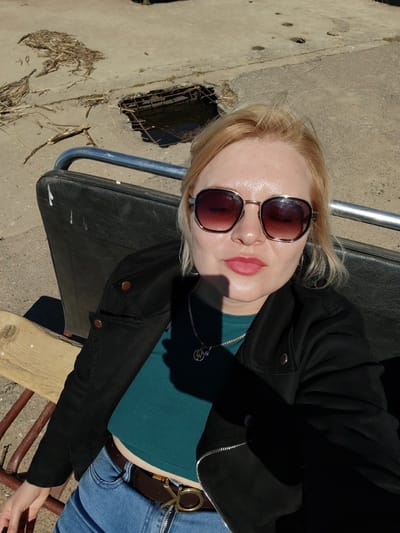 A woman with blonde hair and sunglasses sits on a bench outside. She is wearing a black jacket over a teal crop top and blue jeans with a brown belt. In the background is a concrete surface with a hole filled with water. The location is Икское Устье, Russia.