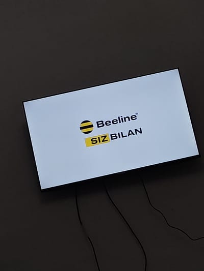 The image shows a television screen displaying a logo that reads "Beeline" with a black and yellow bee icon, and below it, the text "SIZ BILAN" in yellow on a black background. The screen is illuminated white. The television is mounted on a dark gray wall. Two black cables are visible extending from the bottom of the television and disappearing into the darkness of the wall. There are no people or any other discernible objects or details in the scene. The image does not contain any specific information about the city of Gurlan, Uzbekistan.