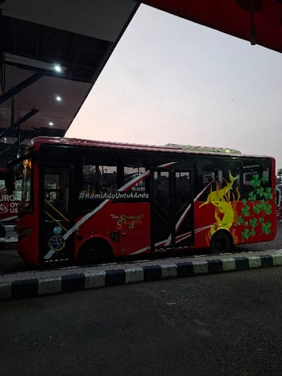 A red and black bus with the words "#KamiAdaUntukAnda" written on the side is parked under a covered area. The bus is in a bus station or bus stop. The bus has a yellow logo of a bird on the side with the words "Turn Smenjal Surabaya" below it. There are green clover leaves along the side of the bus. The bus is parked on a black asphalt surface with white curbs and a black and white tiled area in the foreground. The bus is parked under a metal roof with a few lights visible. The sky is clear and pale blue with the sun setting in the distance. The bus is in the city of Kecamatan Waru, Indonesia (latitude: -7.351, longitude: 112.725).