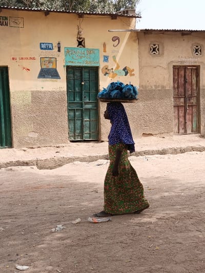 The image portrays an outdoor street scene in Zango, Nigeria, under daylight conditions. A dark-skinned female individual is captured in profile, walking from left to right on a light brown dirt road. She wears a blue patterned head covering and a long, green and orange patterned dress, and appears to be barefoot. Balanced on her head is a dark, circular tray containing multiple irregularly shaped bundles wrapped in blue material.

In the background, two light-colored buildings with corrugated metal roofs are visible. The building on the left features a green double door, secured by a padlock, and its facade is covered with hand-painted advertisements and graphics. These include a red "NO SMOKING" symbol, a handshake logo, an MTN logo with associated phone numbers, a laptop graphic, and the text "ASHRU NA SOMA Communication & Soft Drinks." The building segment to its right has a textured lower wall. Adjacent to this, a second light-colored building presents a distressed dark brown double wooden door. Above this door, text identifying "S.M.G Tailoring" and phone numbers are discernible, flanked by two decorative ventilation blocks. The dirt road in the foreground contains scattered litter, including several discarded plastic bottles. Sparse green foliage is visible above the building roofs.
