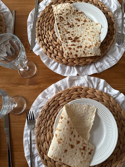 A top-down, medium shot captures a wooden table set for two with plates of matzah. Each place setting includes a woven placemat, a white linen napkin, a white plate, silverware, and a glass. Two clear glasses are partially visible on the left side of the image. The matzah on the plates is flatbread, some pieces folded and others laid flat. The lighting is natural, creating soft shadows. The scene appears to be in a home setting in Starokorsunskaya, Russia.