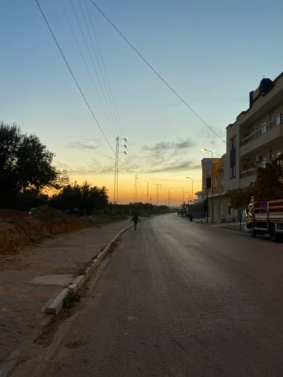 The image depicts a street scene in الغنادة, Tunisia, during sunrise or sunset. The sky displays a gradient from deep orange and yellow at the horizon to light blue overhead, with scattered wispy clouds. A paved asphalt road, appearing somewhat damp, occupies the foreground and curves gently to the right into the midground.
On the left side of the road, an unpaved dirt shoulder with a concrete curb separates the asphalt from elevated mounds of earth and dense green vegetation, including multiple trees and bushes. A series of utility poles, some with cross arms and insulators, are visible along the left side, supporting multiple overhead power lines that extend diagonally across the sky.
On the right side of the road, a row of multi-story buildings, consistent with residential or mixed-use structures, lines the street. These buildings feature balconies and varied architectural details. Several illuminated streetlights are positioned along this side of the road. A parked flatbed truck, featuring a red and blue striped cargo bed, is visible on the far right in the foreground.
One individual, silhouetted against the bright horizon, walks in the middle of the road, away from the viewer, with their arms slightly extended to the sides. A second, smaller figure is discernible further down the road on the right sidewalk near the buildings.