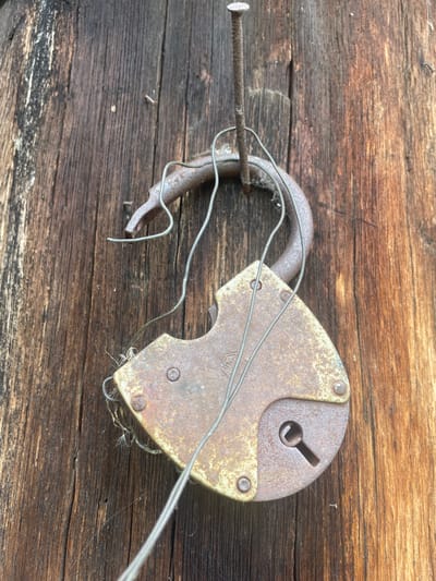 This is a close-up, overhead shot of a rusty, old padlock and a nail attached to a weathered wooden surface. The padlock is partially open, with its shackle bent and intertwined with thin wires. The nail is driven horizontally into the wood, with the padlock's shackle looped around it. The wooden surface appears to be planks, with visible grain and wear, suggesting an outdoor or rustic setting. The lighting is consistent and diffused, indicating it might be daytime, but the time of day is not strongly suggested by any shadows. There is no indication of weather. The scene evokes a sense of age, disuse, and perhaps abandonment. There is no visible text in the image. The location context is given as Валки, Russia.