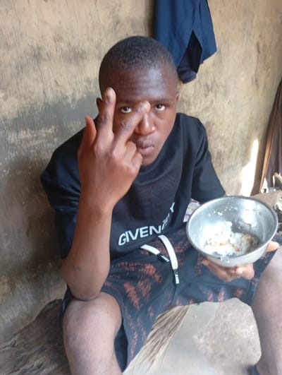 A young man is seated, looking directly at the camera with a neutral expression. He has short, dark hair and dark skin. He is wearing a black short-sleeved t-shirt with the word "GIVENCHY" visible in white letters on the front, and black patterned shorts with white drawstrings.
In his right hand, he holds a small, metallic bowl containing a meal, which appears to be rice with some other food items. His left hand is raised in a gesture where his index and middle fingers are extended, and his ring finger, pinky, and thumb are curled. His extended fingers are positioned near his nose or forehead.
The background consists of a rough, light brown or beige wall, suggesting an indoor or sheltered outdoor setting, consistent with a rural or simple dwelling, possibly in Obwe, Nigeria. In the upper right corner, some dark fabric, likely an item of clothing, hangs against the wall. The lighting is diffused, indicating it is daytime but without direct sunlight.