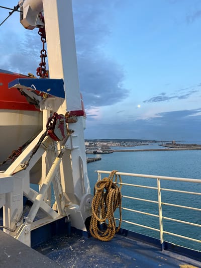 The image depicts a view from the deck of a ship in Civitavecchia, Italy. The city’s harbor is visible in the distance, under a sky with scattered clouds and a visible moon. In the foreground, there is a white railing, and a coiled rope rests against it. Metal structures, painted in white, red, and blue, are part of the ship's machinery. The ship is docked and there are no people visible.