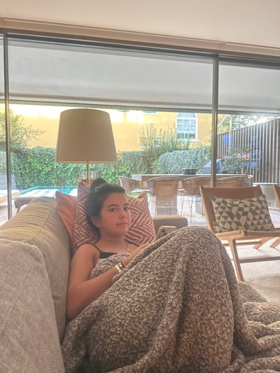 A female subject, appearing adult, is seated on a light grey upholstered couch, partially covered by a textured light brown blanket. She wears a dark top and bracelets on her right wrist, holding a light-cased smartphone beneath the blanket. Her dark hair is pulled back, and she looks directly forward with a neutral expression. Behind her, two decorative pillows are visible. To her left, a floor lamp with a beige drum shade and gold-toned base stands near a large glass window.
Through the window, an outdoor patio features a light-colored wooden deck, a partially visible light-blue swimming pool with a lounge chair, and a dining area with a long table and multiple wicker-style chairs. A wooden leisure chair with a green and white geometric cushion is on the right. A green hedge forms a boundary beyond the patio, backed by a yellow-walled building with white windows. Another building or fence with vertical wooden slats is visible on the far right. The scene depicts an indoor living space looking out onto a private outdoor area during daylight hours.
The precise location within Lordelo do Ouro e Massarelos, Portugal, cannot be determined from the provided image.