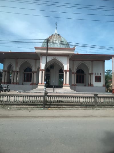The image presents a daytime view of a mosque in RawangItiek, Indonesia, under a clear, bright blue sky. The mosque is a light-colored building, possibly white or cream, adorned with reddish-brown trim and window frames. It features a prominent central dome, which appears translucent or light-toned with a grid pattern, topped by a finial depicting the Islamic crescent and star symbol.

The mosque's facade includes multiple arched entrances and windows, supported by decorative columns. Steps lead up to the entrances. To the right of the mosque, a light-colored, ornate fence with balusters separates the building's grounds from a paved, gray road. A motorcycle is partially visible, parked near one of the mosque's entrances.

Numerous black power lines stretch across the upper part of the sky, originating from a pole that extends vertically along the right side of the mosque. Some green foliage is visible in the background, behind the mosque. The bright lighting indicates it is likely morning or early afternoon. A small, white rectangular sign with dark text, possibly reading "WC" and other illegible characters, can be faintly seen near a lower entrance of the mosque. The overall scene is static, portraying a common street view.