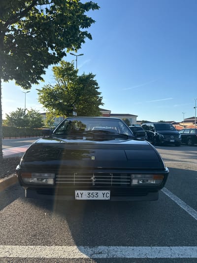 A black Ferrari Mondial is parked in an asphalt lot with white parking lines under a clear blue sky. The car's front end, including the grille and headlights, is prominently featured. Behind the Ferrari, several other vehicles are parked, including a black Mercedes-Benz Vito van and a dark-colored sedan. The background shows buildings and trees. The scene is lit by natural sunlight, casting shadows. The location is the Serravalle Designer Outlet, a shopping mall in Serravalle Scrivia, Italy.