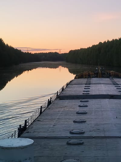The image presents a tranquil scene from the deck of a large vessel, likely a barge or cargo ship, navigating a calm waterway at either sunrise or sunset near Devyatiny, Russia. The viewpoint is from the forward part of the vessel, looking towards the horizon.

In the foreground, the flat, grey deck of the boat dominates the lower right portion of the frame. It features rows of oval-shaped metal hatches or covers. A sturdy metal railing extends along the left edge of the deck, and a white, cylindrical mooring post is partially visible in the bottom left corner.

The tranquil waterway, possibly a river or canal, reflects the soft, warm hues of the sky. A gentle wake trails along the left side of the vessel, indicating movement. Both banks of the waterway are densely covered with dark green forest, creating a lush, natural corridor.

The sky above displays a gradient of soft colors, transitioning from a pale yellow-orange near the horizon to a lighter, almost pinkish hue higher up, characteristic of the golden hour. In the distance, several tall electricity pylons are visible above the treeline on both sides of the waterway, providing a subtle hint of human infrastructure. A small white buoy can also be seen in the water along the left bank.

The overall mood is peaceful and serene. No people are visible in the image.