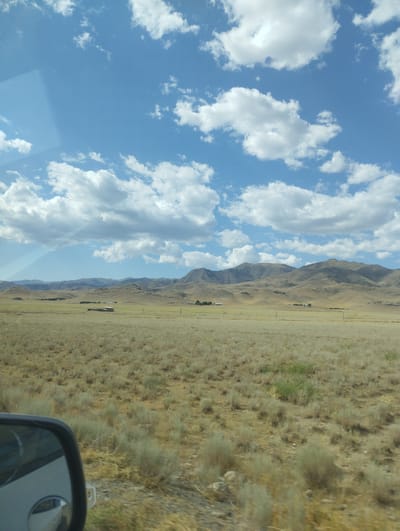 The image depicts a wide, arid landscape under a partly cloudy sky. In the foreground, a field of dry grass and low-lying shrubs stretches towards a range of rolling, barren hills in the background. A small, isolated building with a pitched roof is visible in the middle ground, situated in the center of the frame. The sky is blue with scattered white cumulus clouds, casting shadows on the landscape below. The scene appears to be a rural or undeveloped area. There are no people or vehicles clearly visible in the main part of the image, though a portion of a car's side mirror is reflected in the lower left corner. The overall impression is one of vastness and natural, undeveloped terrain.