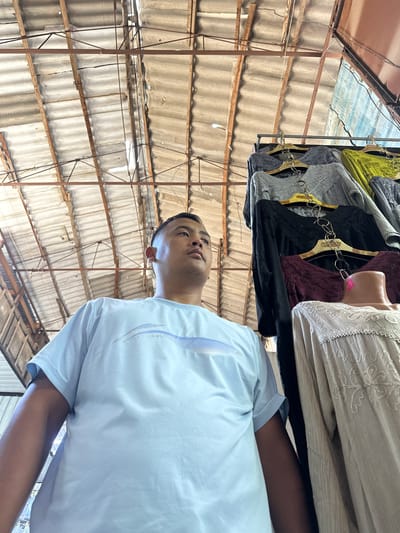 The image depicts an outdoor marketplace setting in Kara-Suu, Kyrgyzstan, characterized by a corrugated metal roof supported by weathered wooden beams and metal struts. In the foreground, a young man wearing a light blue t-shirt with a subtle blue and white stripe design is positioned on the left side of the frame, looking upwards and to the right. To his right, a rack of clothing hangs, featuring several women's tops in various colors: grey, black, burgundy, and lime green. A mannequin or display bust wearing a cream-colored, embroidered garment is also visible on the right. The overall scene suggests a bustling market environment with a focus on textiles.