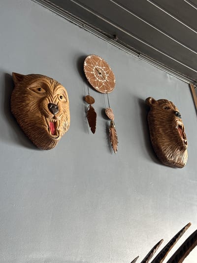 This image depicts a decorative wall display in a room. On a light blue-grey wall, two carved wooden bear heads are mounted, one on the left and one on the right. Both bear heads have their mouths open, revealing carved teeth and red interiors. Centered between the bear heads and slightly above them is a small dreamcatcher. The dreamcatcher is round with a woven pattern and has several small, feather-shaped ornaments hanging from it by thin threads. The ceiling above the wall is made of dark, parallel wooden planks. The room appears to be indoors, possibly in a home or a small business. No people are present in the image.