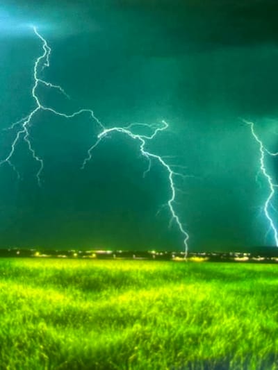 This image depicts a stormy twilight or nighttime scene over a landscape located in Zelenyi Hai, Ukraine. The upper portion of the frame is dominated by a dark, deep teal-green sky, indicative of heavy cloud cover during a thunderstorm. Multiple prominent, bright white-to-light blue lightning bolts are captured across the sky, with at least five distinct, branching strikes visible, illuminating portions of the dark clouds. Along the horizon, a thin, blurred band of diffused yellow and white lights indicates the presence of a distant urban or residential area. The lower half of the image features an expansive field of intensely vibrant green grass, which appears somewhat textured and slightly blurred, suggesting either movement, a long exposure, or digital processing. No people, animals, or specific structures other than the distant lights are visible within the frame. The overall lighting is primarily provided by the lightning flashes and the distant artificial lights, creating a dramatic contrast with the dark sky and luminous green foreground.