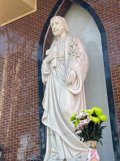 A white marble statue of a male figure, likely religious, stands within a dark arched niche against a brick wall. The figure is depicted from the waist up, holding a cross in its right hand and a bouquet of flowers in its left. The statue has a beard, flowing hair, and wears a draped robe adorned with carved flowers on its shoulder. In the foreground, a vase containing a bouquet of green and pink chrysanthemums is placed at the base of the statue. The brick wall behind the statue is red and appears to be glazed, reflecting light. The niche itself is lined with lighter colored tiles. Above the niche, a wooden soffit with horizontal planks is visible. The overall setting suggests an outdoor or semi-outdoor religious or memorial location, possibly within a church or cemetery. The image was taken in Tuý Sơn, Vietnam.