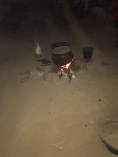The image depicts a dark, likely nighttime, scene showcasing an outdoor cooking arrangement. A large, dark pot with a lid is centrally placed over a bright, open fire, fueled by burning wood. Several rocks are strategically positioned around the fire, serving as a makeshift stove to support the pot. The fire casts an orange glow, illuminating the immediate area. A small animal, resembling a cat or a small dog, is visible on the ground near the fire. To the right of the main cooking area, a smaller, dark bucket or pot is present. The ground beneath is light brown, appearing to be dirt or sand. In the upper right corner, a large, dark cylindrical object, possibly another bucket or a tire, is partially visible. This traditional cooking method suggests a rural or undeveloped setting, aligning with the provided location context of Ago-Abo, Nigeria. No people are visible, and there is no discernible text.