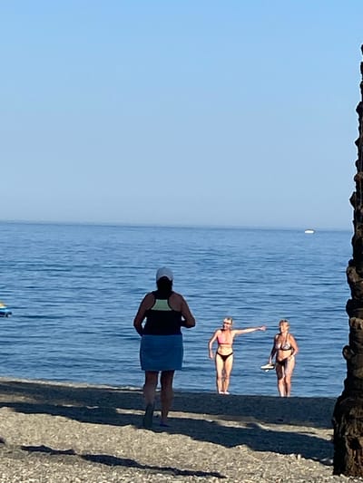The image shows three women on a sunny day by the sea. Two women in bikinis are standing on a concrete pier, looking out at the water. One woman is wearing a pink bikini and the other a black and white patterned bikini. A third woman, viewed from the back, is wearing a dark top, a blue skirt, and a white hat, standing on the same pier. The sea is blue with visible waves. The sky is clear and light blue, suggesting good weather. A rough, rocky structure is visible at the top of the image, and a textured, grey concrete surface occupies the right side. The scene appears to be a recreational area, likely a beach or a promenade in Marbella, Spain, given the context. The time of day is likely daytime, given the bright sunlight casting shadows. There is no visible text in the image.