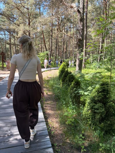 In Morskoe, Russia, a woman with blonde hair pulled back walks along a wooden boardwalk in a pine forest. She is wearing a light-colored cropped t-shirt, dark brown baggy pants, and white sneakers. A crossbody bag with a patterned strap is slung over her shoulder. She holds a mobile phone in her right hand. Behind her, several people are walking on the boardwalk, some in casual attire, including a man in a white shirt and beige pants, a man in a blue t-shirt and dark shorts, and a person in a white dress. The boardwalk is bordered by dense green vegetation, including manicured evergreen shrubs and tall pine trees with a clear blue sky visible through their branches. The ground is covered with pine needles and other natural debris. The lighting suggests it is daytime with sunlight filtering through the trees.