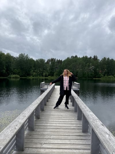 A woman is standing on a wooden pier that extends into a calm, dark lake. She is wearing a black quilted jacket over a pink and white checkered shirt and black pants. She has her left arm outstretched and is looking to her left with her hair blowing. The pier is made of weathered grey wood with railings. Behind the lake, there is a dense forest of green trees, and in the distance, houses are visible amongst the trees. The sky is overcast with grey clouds. This image was taken in the city of Mars, Russia.