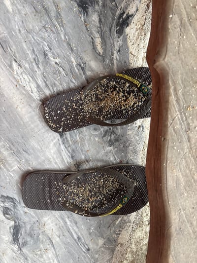 A pair of dark brown Havaianas brand flip-flops, heavily coated with light-colored sand and small shell fragments, are depicted resting on a grey and white marbled surface. The flip-flops are positioned vertically, with the yellow "Havaianas" logo and a Brazilian flag emblem visible on the outer strap of each. The surrounding marbled surface is also scattered with sand particles. To the right, a distinct vertical boundary separates the marbled surface from a lighter, coarser, sandy-textured ground, partially bordered by a vertical, light brown wooden post or natural structure. The scene is consistent with a coastal or beach-proximate area on Koh Phangan, Thailand. No individuals are present.