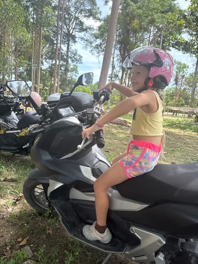 A young blonde girl wearing a pink helmet, a yellow tank top, and tie-dye shorts sits on a black and silver scooter. She is looking to her right with a smile. Her hands are on the scooter's handlebars. Another scooter with a black and yellow design is parked behind her. The scooters are parked on a dirt path surrounded by trees and vegetation. The location is Baan Harn, Thailand.