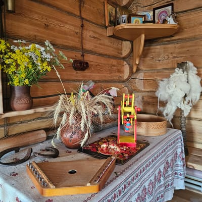 The image depicts an interior scene with a strong rustic and traditional Russian aesthetic. The setting is a room with wooden walls and floor, suggesting a rural dwelling or a historical exhibit. Various objects are arranged in a seemingly domestic yet curated manner.

Prominently featured are religious icons, stacked on a wooden shelf on the left. A wooden spindle with fluffy white wool is visible hanging from the wall, hinting at textile crafting. In the center, a colorful wooden toy airplane is placed on a decorative tray.

On the right side of the image, a table is covered with a cloth featuring a red and white floral pattern. On this table rests a stringed musical instrument, possibly a gusli or zither, and a decorative wooden box. A bouquet of wildflowers, including daisies and yellow flowers, is arranged in a rustic clay jug. Stalks of wheat are also present, adding to the harvest or rural theme. Two horseshoes are placed on the cloth, suggesting a possible good luck charm or connection to horses. A wooden rolling pin lies on its side near the jug.

The lighting is soft, and there are no clear indicators of time of day or weather. The overall impression is one of a preserved traditional environment, possibly a museum display or a thoughtfully decorated home. There is no visible text in the image.