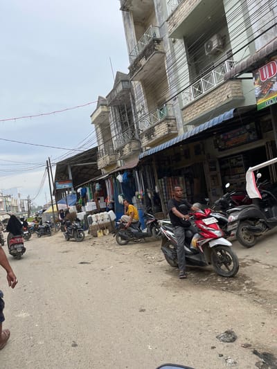 In Kota Kuala Simpang, Indonesia, a street scene unfolds with a focus on a dirt road lined with market stalls and buildings. The sky is overcast. In the foreground, a person wearing sandals and shorts is visible, their hand with a ring on their index finger reaching out of frame. To the left, a woman in a black hijab rides a red scooter. Further down the road, several other scooters are parked or being ridden, including a red and white Honda scooter with a man in a black shirt and grey pants. Behind the man on the Honda, a black scooter and a dark grey scooter are parked. To the right, men are seated or standing near market stalls that display various goods, including stacks of white containers, baskets with tools, and rolls of fabric. The buildings have multiple stories with balconies, and many electrical wires crisscross above the street. A sign with Indonesian text is visible on one of the stalls. The overall environment appears to be a busy marketplace with a mix of commerce and daily life.