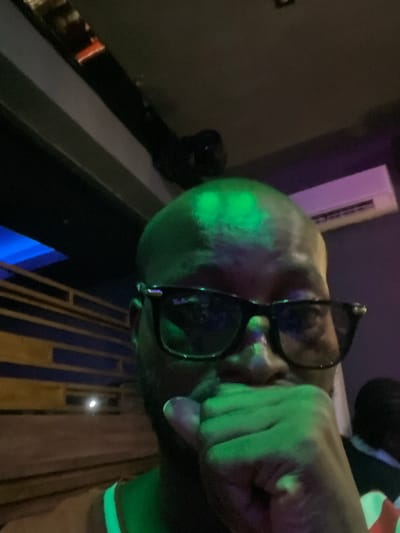 A person with a bald head and dark-framed sunglasses is illuminated by a green light. The individual's hand is raised near their face. The background shows parts of a building or structure with blue and yellow elements. The image appears to be taken at night or in a dimly lit environment, but the location in the city of Rubuchi, Nigeria, cannot be determined from the visual information provided.