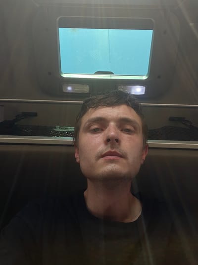 A close-up, low-angle selfie captures a young adult male with light skin, freckles, and short brown hair looking directly at the camera. He is wearing a black t-shirt and appears to be in an enclosed space with a light-colored ceiling and a large, rectangular skylight above. The environment suggests the interior of a vehicle, possibly a train or bus, given the overhead storage nets visible in the background. The lighting is somewhat dim, with a bright overhead light to the right of the skylight and natural light filtering through the skylight, creating some glare and highlights on the subject's face. The background is out of focus, but appears to be dark upholstery. There is no indication of the specific location in Brachterbeek, Netherlands, in the image itself.